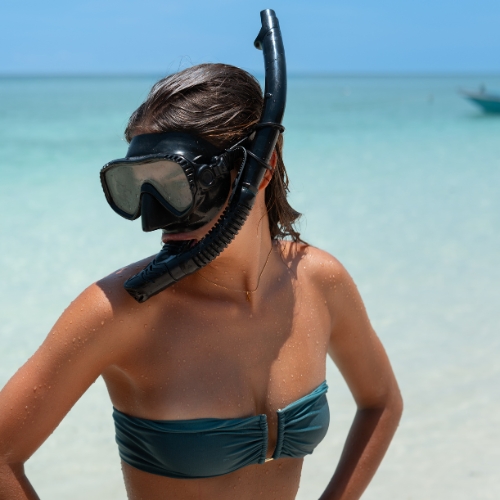 A woman in a stylish blue bikini and black snorkel mask prepares for a snorkeling adventure in the crystal-clear waters of Lombok, with a serene tropical beach in the background.